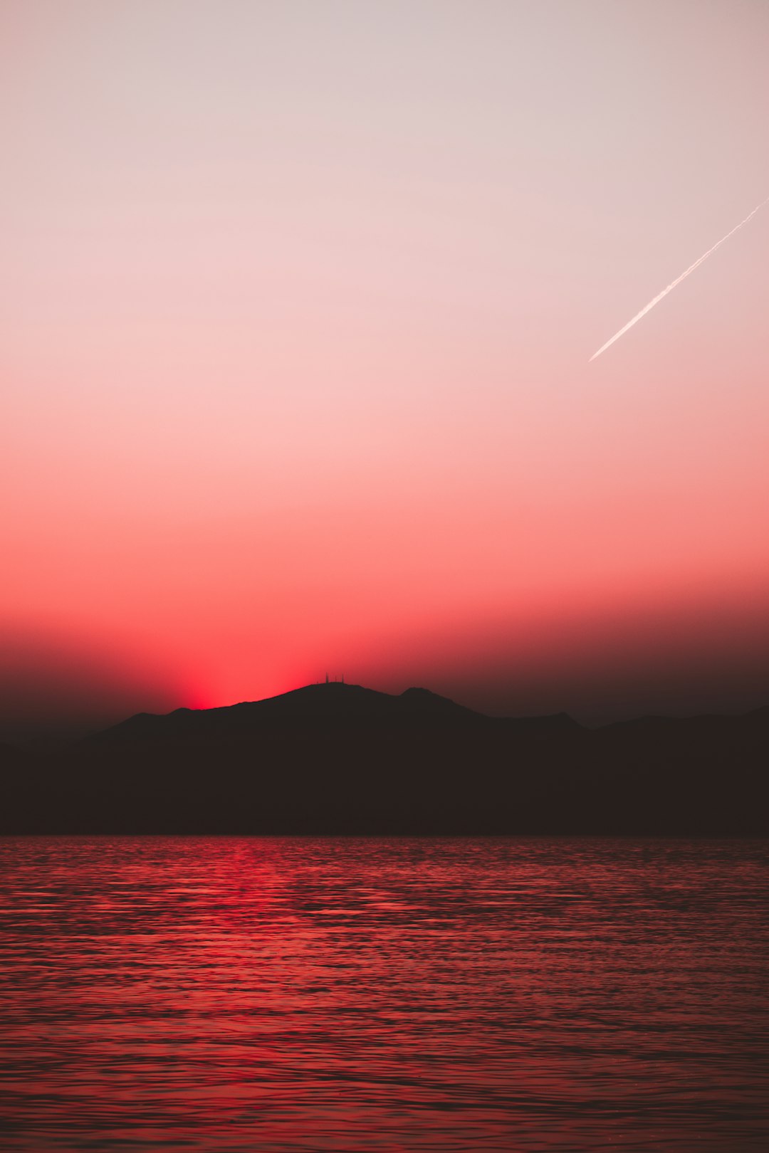 Red sky, distant islands and mountains, sunset, plane trail, calm sea surface, simple composition, minimalist style, wideangle lens, distant view, silhouette effect, soft light, tranquil atmosphere. in the style of minimalist. –ar 85:128