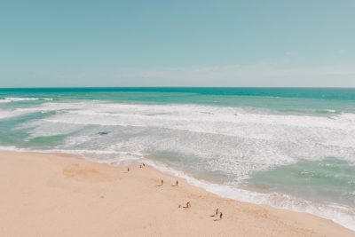 photograph of A beautiful beach with clear blue water and golden sand, people playing in the waves, drone view, wide angle lens, cinematic, minimalism, simple, elegant, relaxing, tranquil, light green and white, sunny day, --ar 128:85