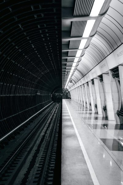 A black and white photo of the interior of an empty modern underground station, with train tracks visible on one side. The walls feature clean lines and geometric shapes in shades of gray or metallic tones. A few people can be seen walking along the platform. In the background is another subway line leading to downtown Moscow. Shot in the style of photographer [Miki Asai](https://goo.gl/search?artist%20Miki%20Asai) using a Leica camera. --ar 85:128