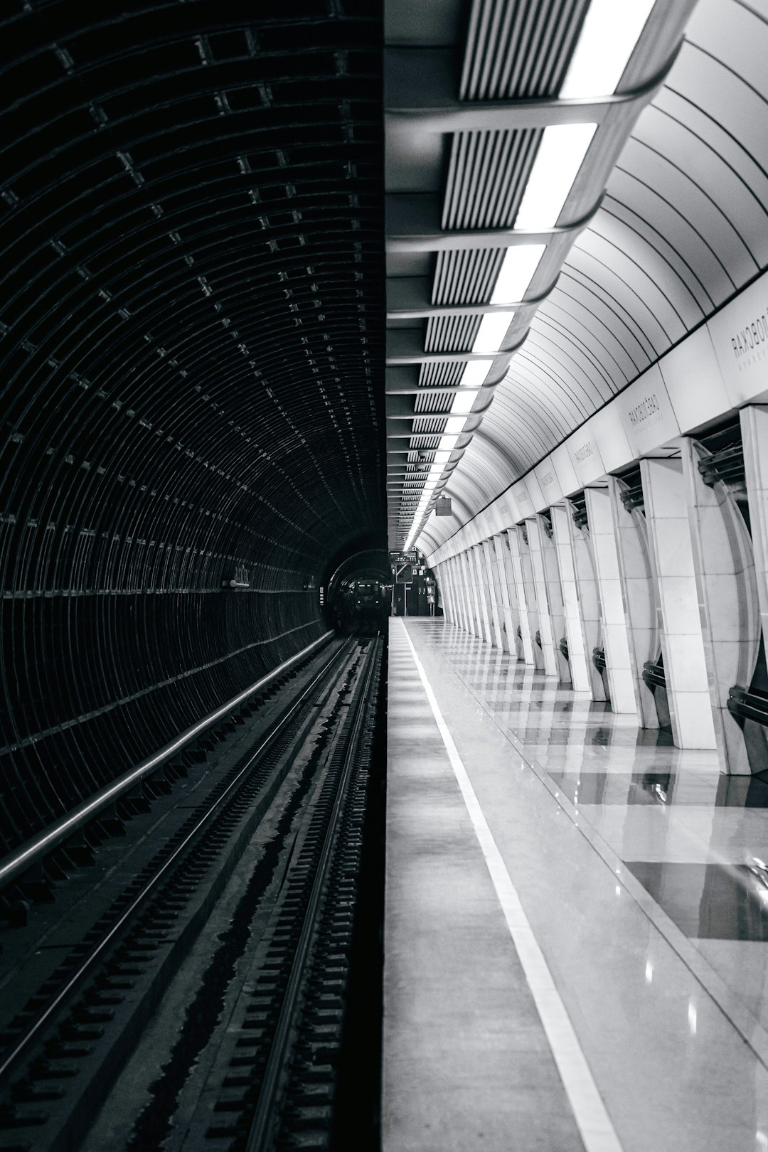 A black and white photo of the interior of an empty modern underground station, with train tracks visible on one side. The walls feature clean lines and geometric shapes in shades of gray or metallic tones. A few people can be seen walking along the platform. In the background is another subway line leading to downtown Moscow. Shot in the style of photographer [Miki Asai](https://goo.gl/search?artist%20Miki%20Asai) using a Leica camera. –ar 85:128