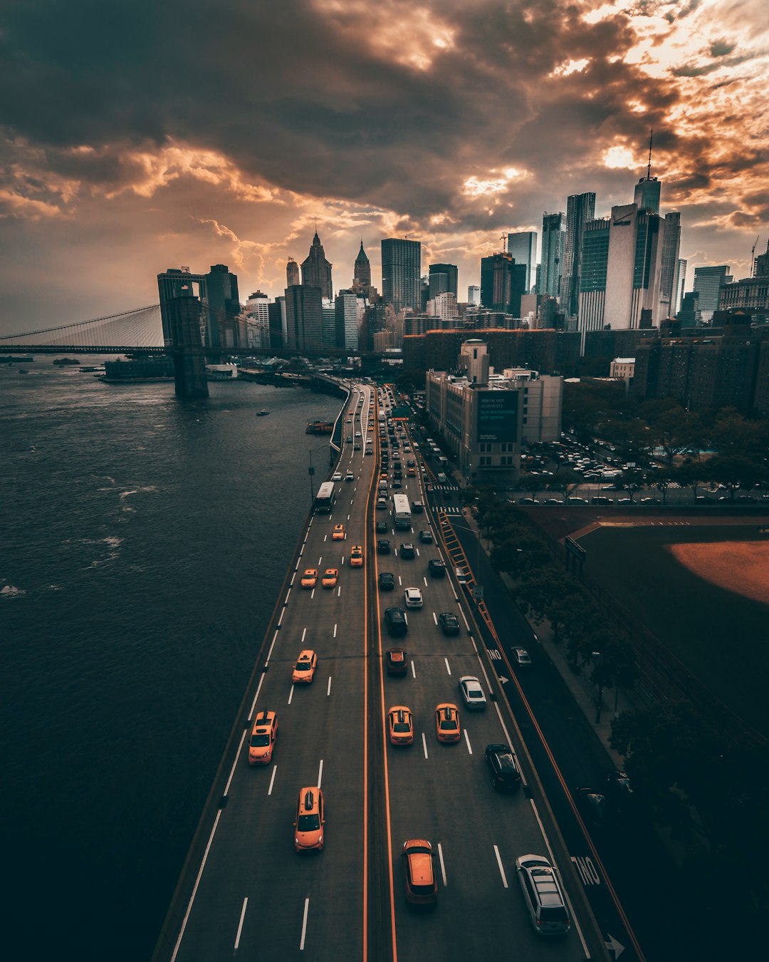 drone photo of New York City, bridge and road with cars, skyline in the background, moody sky, orange lights from city buildings, cars on highway, buildings in distance, –ar 51:64