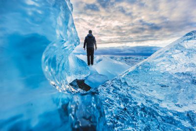 A man stands on an ice wall, surrounded by blue crystal clear snow and ice in Iceland's vast landscape. The sky is cloudy with some sunlight shining through the clouds. He wears warm  to stay covered from the cold weather. Captured using a Canon EOS R5 camera with a wide-angle lens at an f/8 aperture setting. A low angle perspective view of his figure against the majestic scenery in the style of landscape photography. --ar 128:85