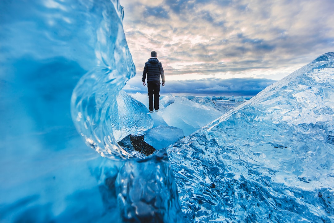 A man stands on an ice wall, surrounded by blue crystal clear snow and ice in Iceland’s vast landscape. The sky is cloudy with some sunlight shining through the clouds. He wears warm  to stay covered from the cold weather. Captured using a Canon EOS R5 camera with a wide-angle lens at an f/8 aperture setting. A low angle perspective view of his figure against the majestic scenery in the style of landscape photography. –ar 128:85