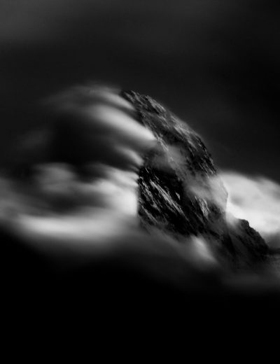 A black and white photograph of an isolated mountain peak, surrounded by swirling mist that creates the illusion of water flowing down its side. The mountain is in sharp focus against a blurred background of misty clouds. Shot with Nikon D850 for a dramatic contrast between light and dark areas. Soft lighting enhances the ethereal atmosphere. --ar 49:64