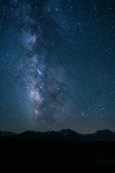 A breathtaking view of the Milky Way galaxy illuminates the dark night sky with stars and distant mountains below. Captured in the style of a professional photographer with high-resolution and sharp photography equipment. The composition is centered on the starry backdrop with the silhouette of mountain peaks adding depth to the scene. High contrast lighting highlights the celestial beauty against the black background. A wide-angle lens provides a full panoramic view. --ar 85:128
