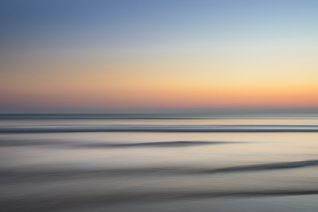 A serene beach at sunset, with gentle waves and soft pastel colors in the sky, captured through long exposure photography. The focus is on capturing motion blur of water ripples against the horizon line, creating an abstract expressionist style that emphasizes fluidity and movement. This scene embodies tranquility and serenity. –ar 128:85