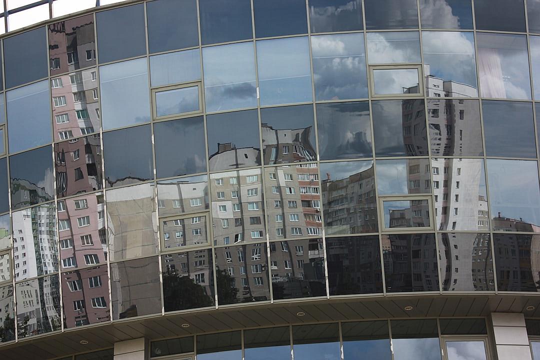 In the city of Minsk, there is an office building with glass walls that reflect other buildings and clouds. The reflection on one wall shows part of another multistory apartment block, while another window reflects residential blocks in different parts of town. This photo was taken from outside using a wideangle lens to capture both the architecture inside and around it.