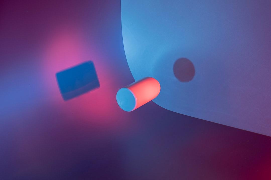 Minimalist composition of two contrasting shapes, one blue and the other pink with a glowing red light source. The neon tube is placed on top of an abstract white background, casting soft shadows on its surroundings. In the foreground, there’s a small square object emitting a subtle glow. Captured from above in a studio setting, this scene creates a visually striking contrast between cool blues and warm pinks, evoking a sense of modernity and minimalism.