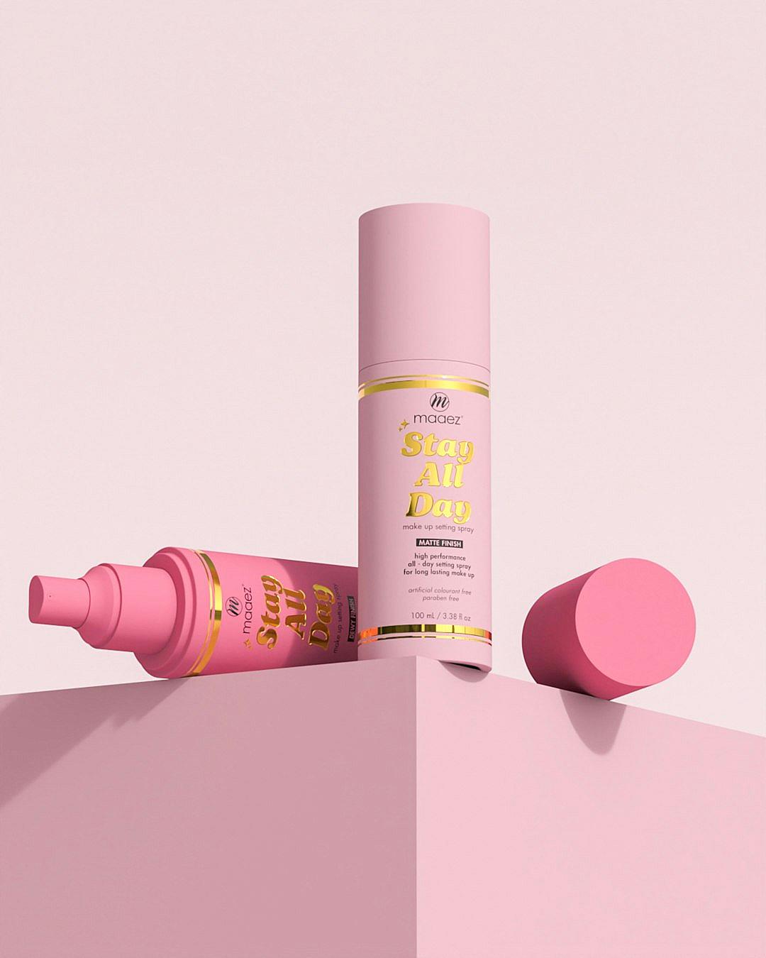 A pink bottle of body spray with a gold cap and a “Stay all Day” label, a pink cylinder shaped box on the side with the brand name in M dee size font, on a light pastel background, product photography in the style of M dee.