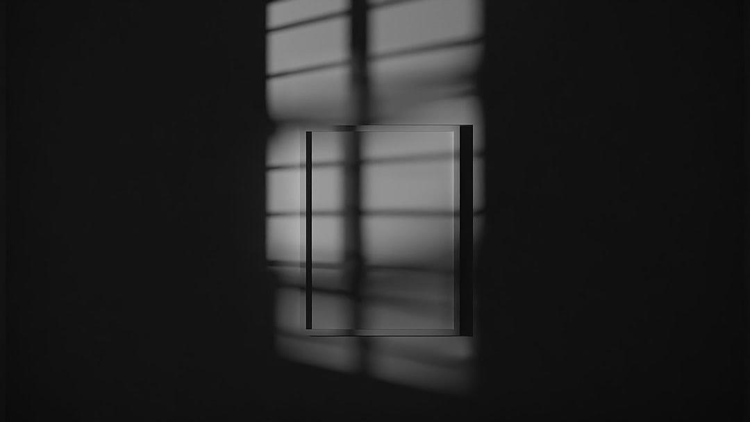 A black and white image of an empty rectangle frame on the wall, with subtle shadows from window panes casting across it, creating a minimalistic yet striking effect. The background is dark grey to accentuate the contrast between light and shadow.