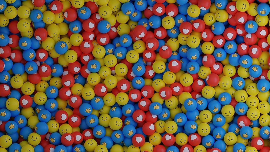 A background of red, blue and yellow balls with smiley faces on them, some have white hearts or thumbs up on their face,