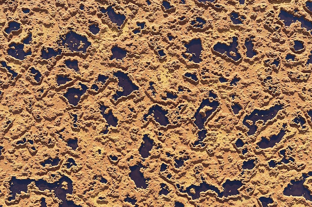 a top down view of the surface texture of earth, with small blue holes in it. The surface is yellowish orange and textured like desert sandstone.