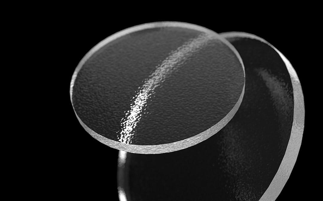 A black and white photo shows two circular glass lenses, one on top and one below. The lens is on its side with some light reflecting off it. There is a vertical line between them that has small sand particles floating inside it. It looks like a closeup shot against a black background.