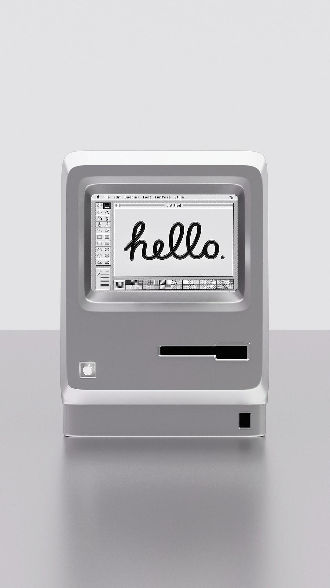 A minimalist gray and white Apple macintosh computer from the first version, with “hello” written on its screen in black cursive text. The background is plain light grey.