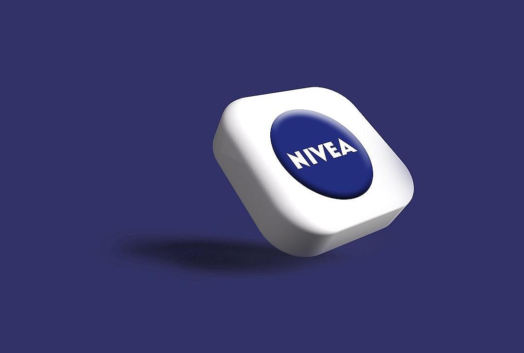 Nivea’s white and blue logo is displayed on the square button, set against an indigo background. The design incorporates clean lines with sharp edges to create a modern aesthetic. A soft shadow falls from above, adding depth and dimensionality to the scene.