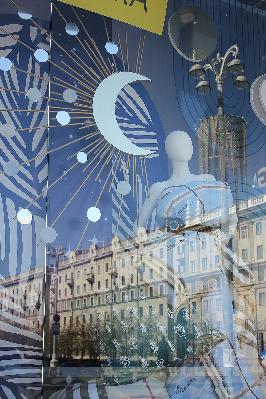 art deco window display in St Petersburg, mannequins wearing a white dress with a blue moon and sun motif print, art nouveau buildings, reflections of the sky on the glass,