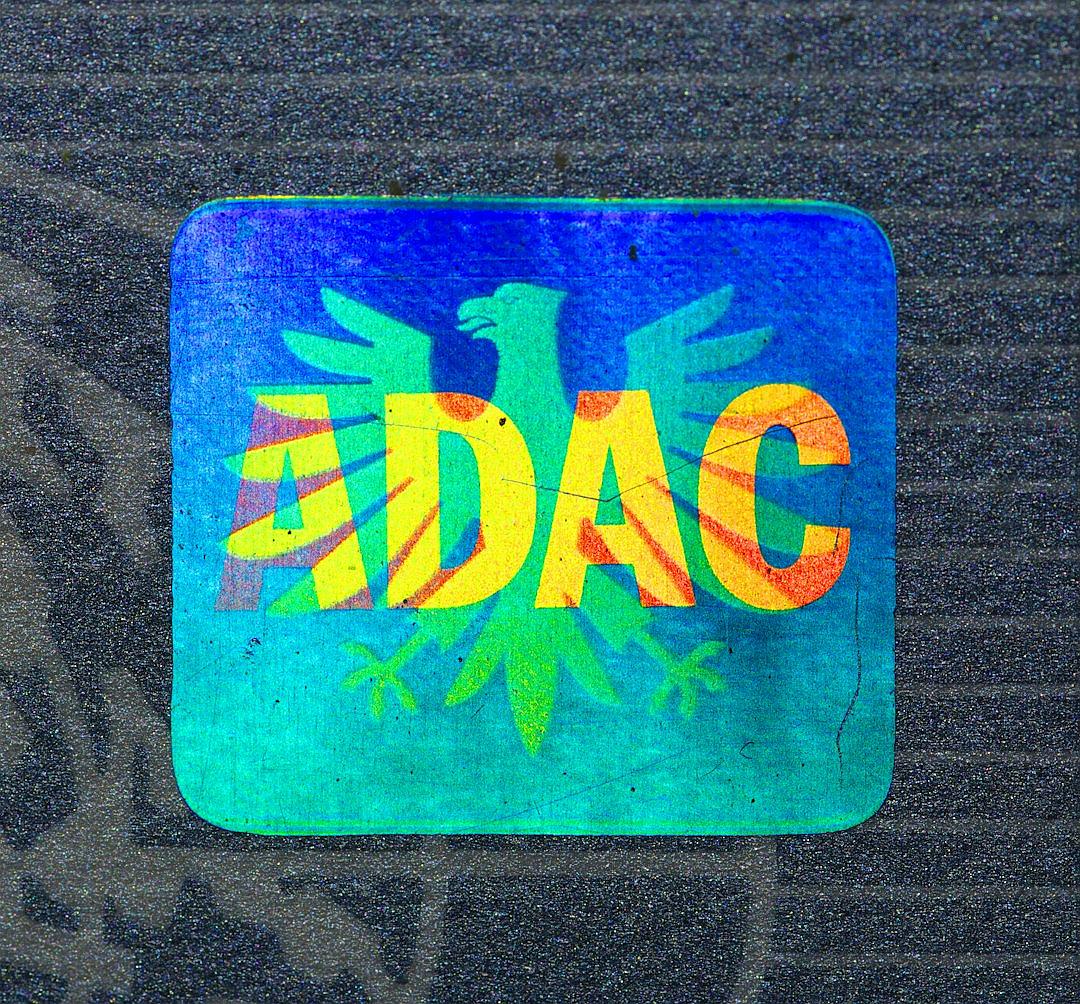 Holographic sticker with the word “ADAC” and an eagle on it, in blue, green, yellow and orange colors, background is gray asphalt.