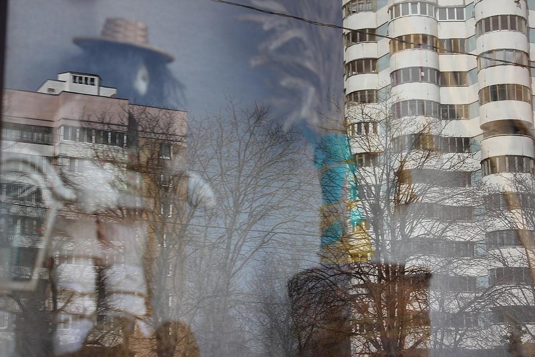 Photograph of Soviet apartment buildings in the city, with trees and people reflected on it. The reflection is distorted by glass windows, creating an abstract urban landscape. In one window there’s blue and yellow paint, adding to its surreal atmosphere.