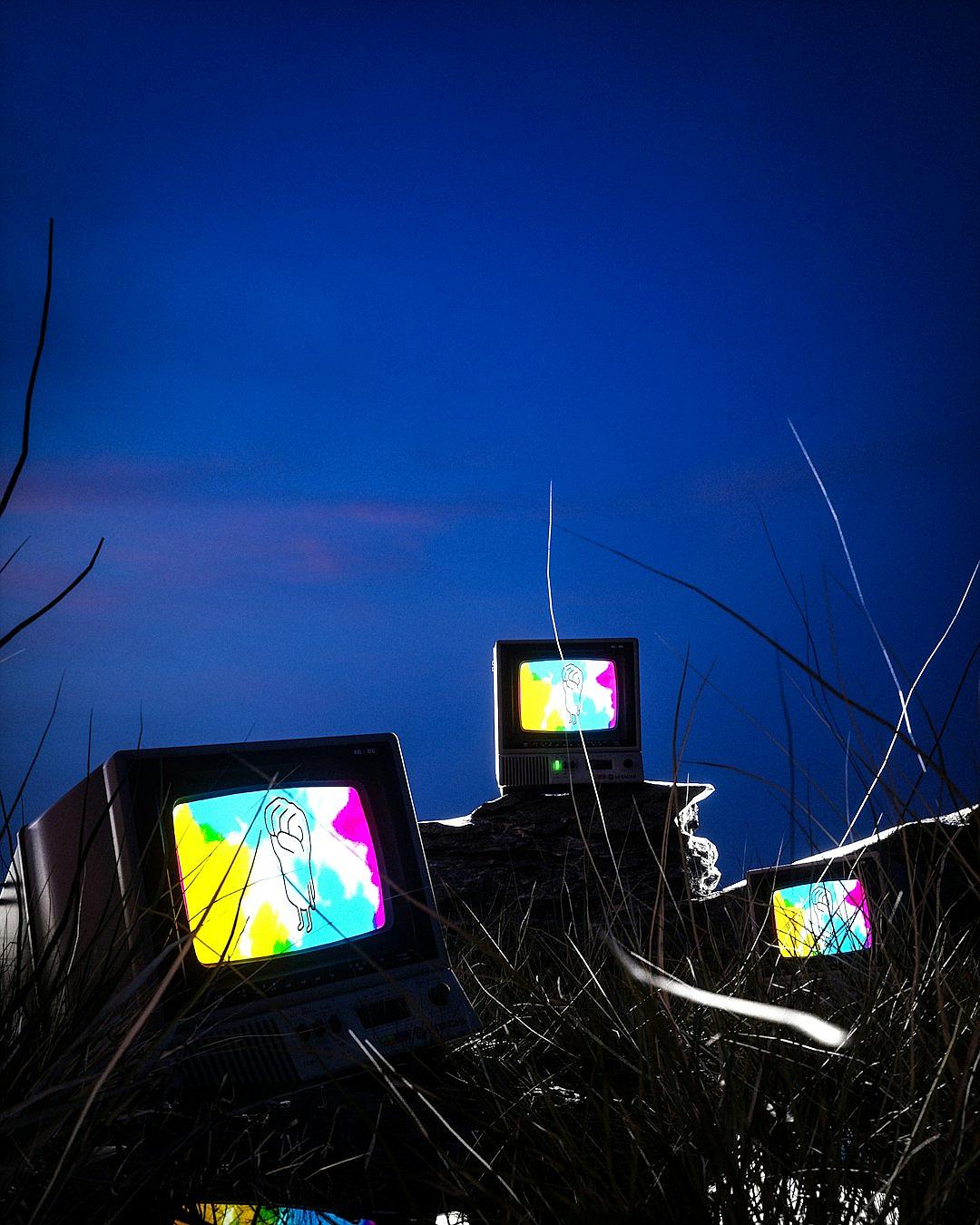 A photograph of three old computer monitors with colourful glitchy screens placed on top of tall grass, in the background is an open dark blue sky at night time from a low angle shot. The photograph was taken in the style of Jimmy Marble and [Bill Henson](https://goo.gl/search?artist%20Bill%20Henson), with no lighting on the ground below.