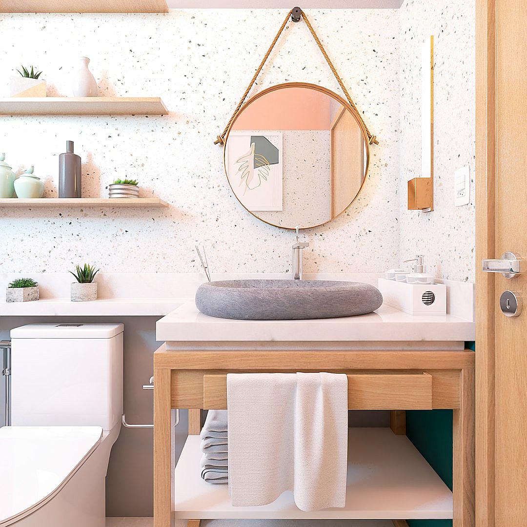 A modern bathroom with white walls, pale pink and grey terrazzo tiles on the floor, pastel green accents, a hanging mirror above an oak vanity that has a round concrete sink, and shelves holding cute decorations. The space is well lit by natural light from outside, creating a warm atmosphere. A towel rack hangs next to it for beach style decor. There’s also a toilet in one corner of the room.