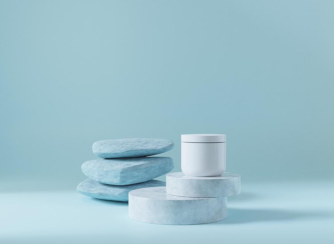 A product photograph of an unbranded white cosmetic jar placed on three stacked light blue stones, against a pastel baby blue background. The scene is captured from the front perspective with soft lighting that highlights the glossy texture and sleek design of both the stone and container. This composition creates a sense of simplicity and elegance, emphasizing the clean lines and modern aesthetic of each element in the setting.