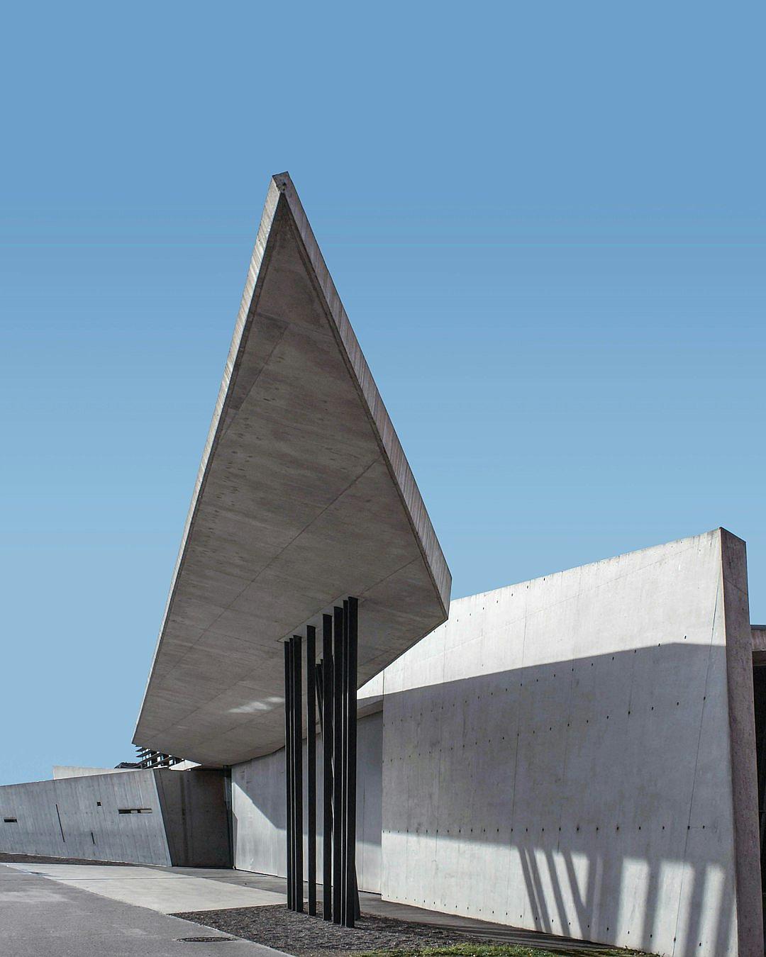 Photograph of the entrance to an architectural museum building with concrete walls and a large triangle shaped roof, surrounded by black metal columns in Bavaria, Germany designed in the style of [Zaha Hadid](https://goo.gl/search?artist%20Zaha%20Hadid).