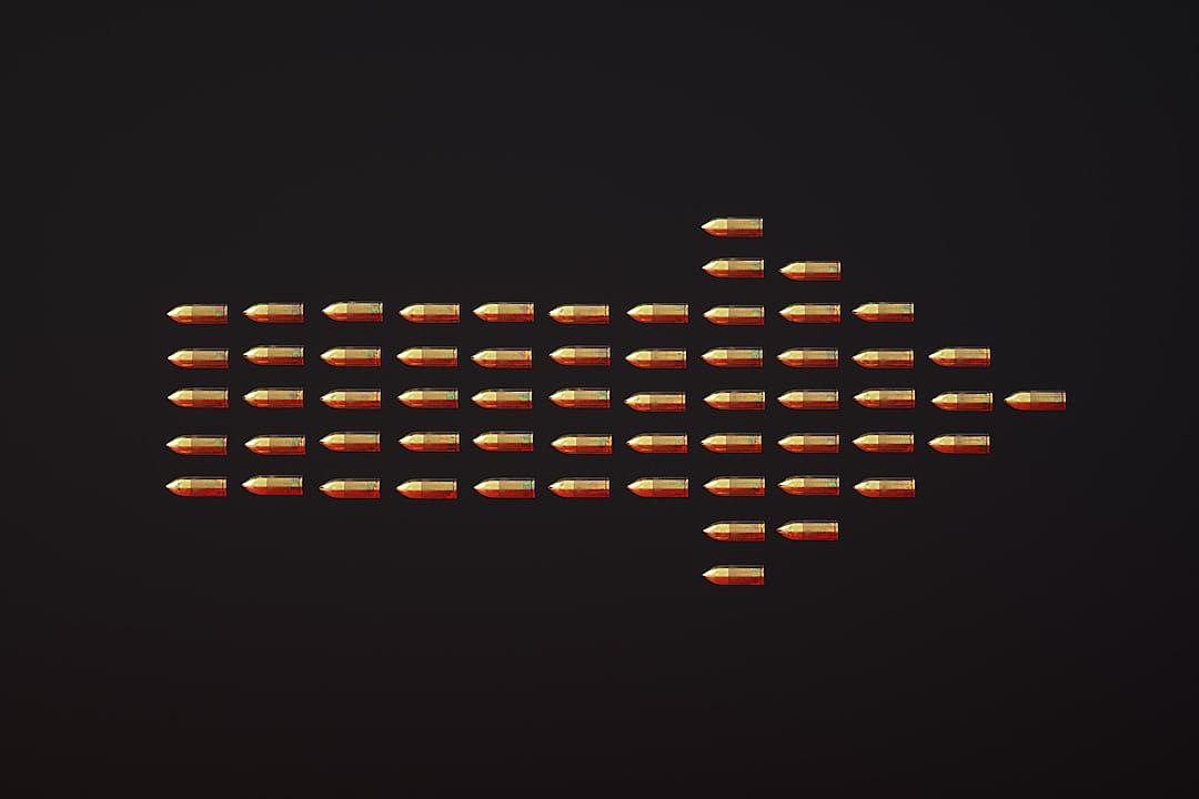 A flat lay of small bullets is arranged in the shape of an upward pointing arrow on a black background. The focus is sharp and detailed, capturing every tiny detail with precision. This composition evokes nostalgia for retro video games while adding elements that highlight modernity through its digital presentation.