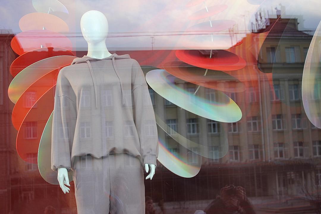 A mannequin in a grey hoodie and trousers, a big red white and orange glass window with a reflection of Russian buildings in the background, colorful large abstract shapes floating around, a depth blur effect, a summer day, a photo taken from outside.