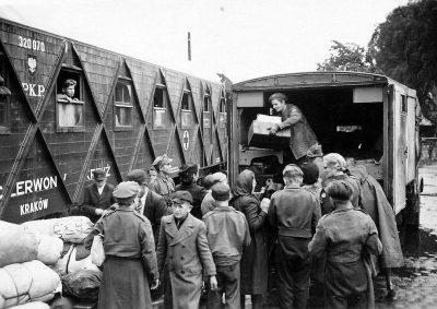 A black and white photograph of an old army cargo train with triangle shaped doors, filled to the brim with soldiers unloading supplies from inside onto people standing in line next outside the door, "Z extracts" written on one side of each box being roadie by some young handsome men wearing overalls and caps holding boxes full of food for Polish children during World War II. It is set at a military base near bending forward railway station with blurred background.