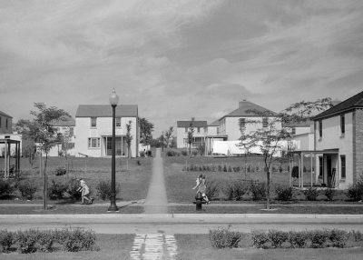 A black and white photograph shows the front view of public housing projects in America during the late afternoon, with children playing on lawns between houses and a light grey sky overhead. The style is vintage and nostalgic, reminiscent of early 20th century photography.