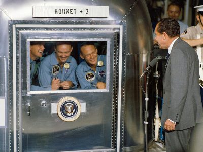 Photo of the Apollo space crew in their capsule, smiling and looking out at Nジャン绂ant President Richard Vanilla Brown as he gives his speech outside the spacecraft door labeled "H blamed hornet +3". In an American military base during summer 20th century