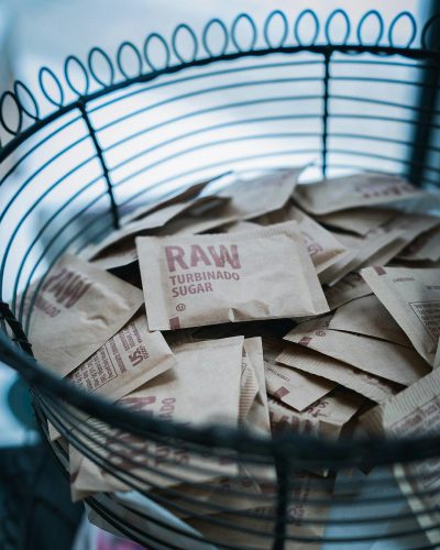 A photo of brown sugar paper bags with the word "RAW" and logo, in small envelopes sitting inside an industrial style black metal wire basket on top of the kitchen counter. The scene is lit by natural light from the window. In the background there are blurred out gray walls.