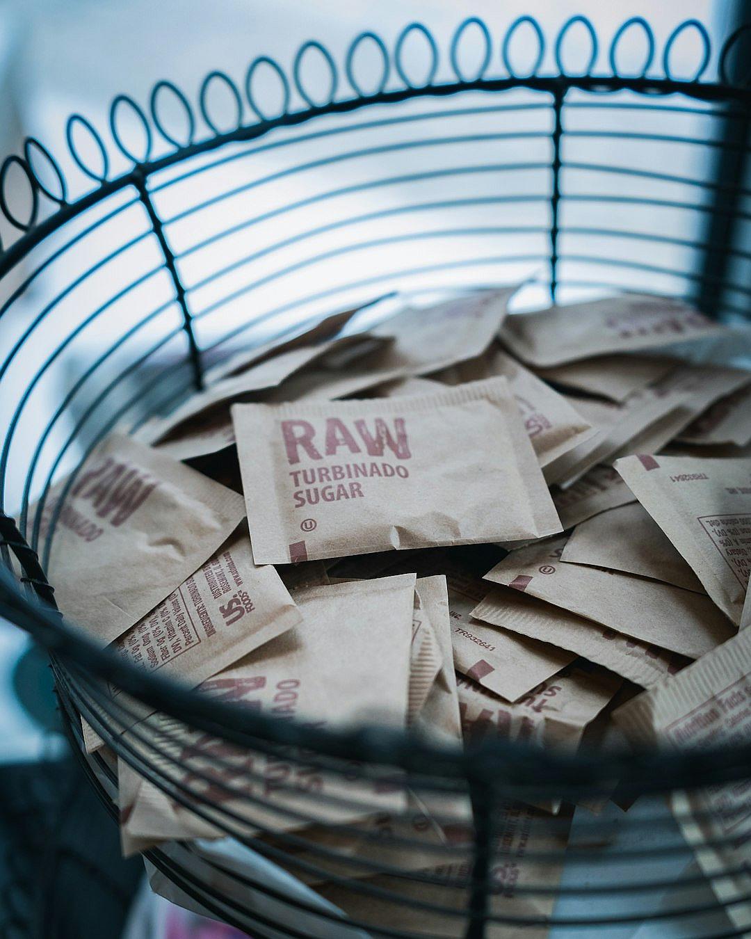 A photo of brown sugar paper bags with the word “RAW” and logo, in small envelopes sitting inside an industrial style black metal wire basket on top of the kitchen counter. The scene is lit by natural light from the window. In the background there are blurred out gray walls.