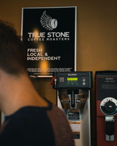 A photo of the "True Stone Coffee Ro vertically and with BUNN coffee machine in front, a large poster on wall behind saying F inflated text that says "FTrump's Re code countryside to urban" A man standing at an industrial style stainless steel espresso station bar with black vinyl lettering that reads "COFFEE workbook cover design has small white phoenix symbol above main title, modern sleek design. In background is a sign reading '‘characters from Marvel’s avengers'’ , vibrant colors, depth of field, Fujifilm GFX 50S