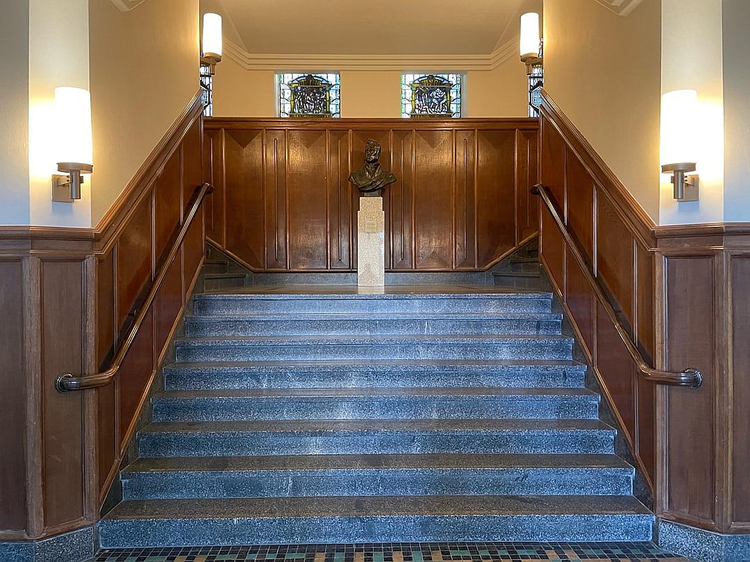 A staircase leads to the second floor of an old university building with wooden panels and blue carpet. Two wall sconces are on each side of the staircase. A small statue is placed at one end of the stairs. The walls have brown wood paneling. There is no door or window in front of you. Stained glass windows line up above in the style of stained glass windows.