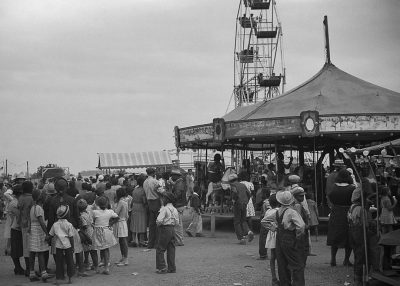 A black and white photograph of an old carnival, with people waiting in line for rides, children playing on the ground near carousels, under a gray sky. The scene is captured from behind in the style of someone standing at a distance, focusing on the crowd's activity. It captures elements like tents, roller coasters, and other amusement park features. Taken during midday light, the photo has a vintage feel to it, reminiscent of classic photography techniques.