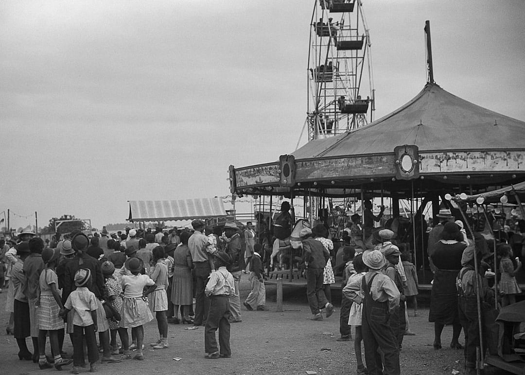 A black and white photograph of an old carnival, with people waiting in line for rides, children playing on the ground near carousels, under a gray sky. The scene is captured from behind in the style of someone standing at a distance, focusing on the crowd’s activity. It captures elements like tents, roller coasters, and other amusement park features. Taken during midday light, the photo has a vintage feel to it, reminiscent of classic photography techniques.