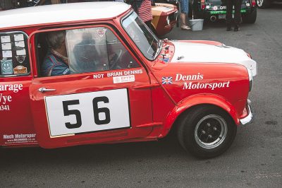 A red and white mini race car with the number "56" on it, which is part of an old British racing event called Japanime int trianglular rally 2018. The driver's name written in English text says not Blanch."Dasily", while another line reads "andrion & dennis". On one side there’s also writing that reads 'The garage', 'mech brain' , '347' and ‘M deaf sport’. In background there are other cars and people.