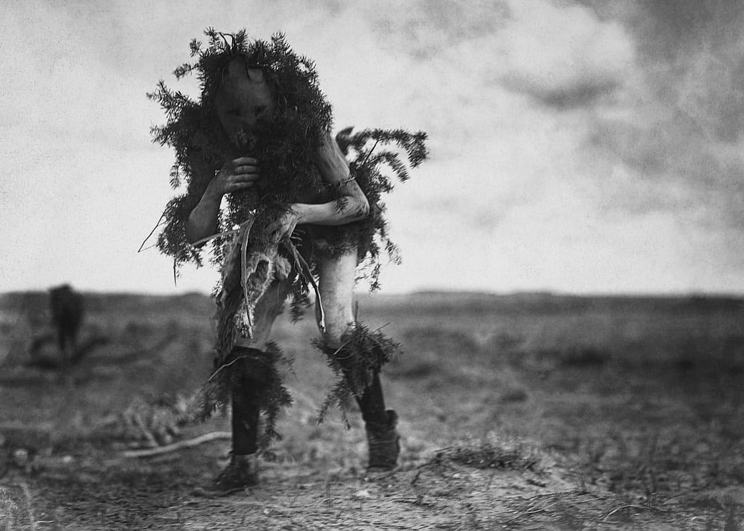 A man covered in grass and twigs dances on the field, in an old photo in black and white style.