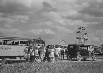 Black and white photo of people getting on the school bus at a fair in Texas. 30's style vintage cars and a ferris wheel are in the background with black clouds overhead. "Valor augusta!" is written on the side of an old-fashioned school bus. A group is standing around and children are laughing in the rural scene. It has the style of vintage photography.