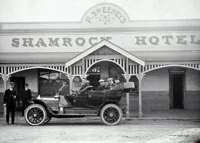 Black and white photo of the front entrance to "S.J.Seven's" hotel in Australia, with people standing outside wearing small leprechaun hats on top of their heads. A black car is parked outside, and there’s an Irish man sitting inside dressed in the style of Irish, with his family around him. The sign above reads 'Shamrock Hotel'.