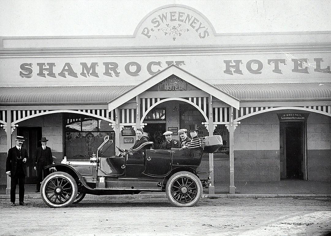 Black and white photo of the front entrance to “S.J.Seven’s” hotel in Australia, with people standing outside wearing small leprechaun hats on top of their heads. A black car is parked outside, and there’s an Irish man sitting inside dressed in the style of Irish, with his family around him. The sign above reads ‘Shamrock Hotel’.