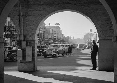 A black and white photo of an archway in downtown glamour, with cars from the 1950s parked on both sides of the street, and people walking around. The photograph was taken in the style of [John Lurie](https://goo.gl/search?artist%20John%20Lurie). It is a sunny day, with sunlight shining through the arched opening onto the road below. A man stands at one end wearing a dark suit jacket and hat looking towards the camera. On his right side is another person dressed in a long coat standing near a car door. They have their backs to each other.