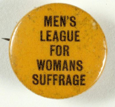 A vintage pinback button with the words "MEN'S LEAGUE FOR WOMAN SUFFRAGE" on it, written in black ink against an isolated white background. The badge is yellow and has wear marks from being worn over time. It's designed to look like something that would have been used during early Women’s Suffrage movement.