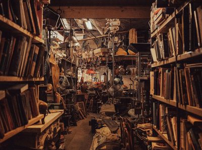 a photo of an old messy bookshop filled with books, trinkets and knickknacks. It's dark inside the shop but there is light coming from above in some places. There’s lots of antique objects on shelves and hanging from ceiling. The atmosphere feels like it would be at night time. In one corner of the room we can see large boxes stacked up covered by black plastic sheeting. At dusk. Shot taken on Nikon Z9