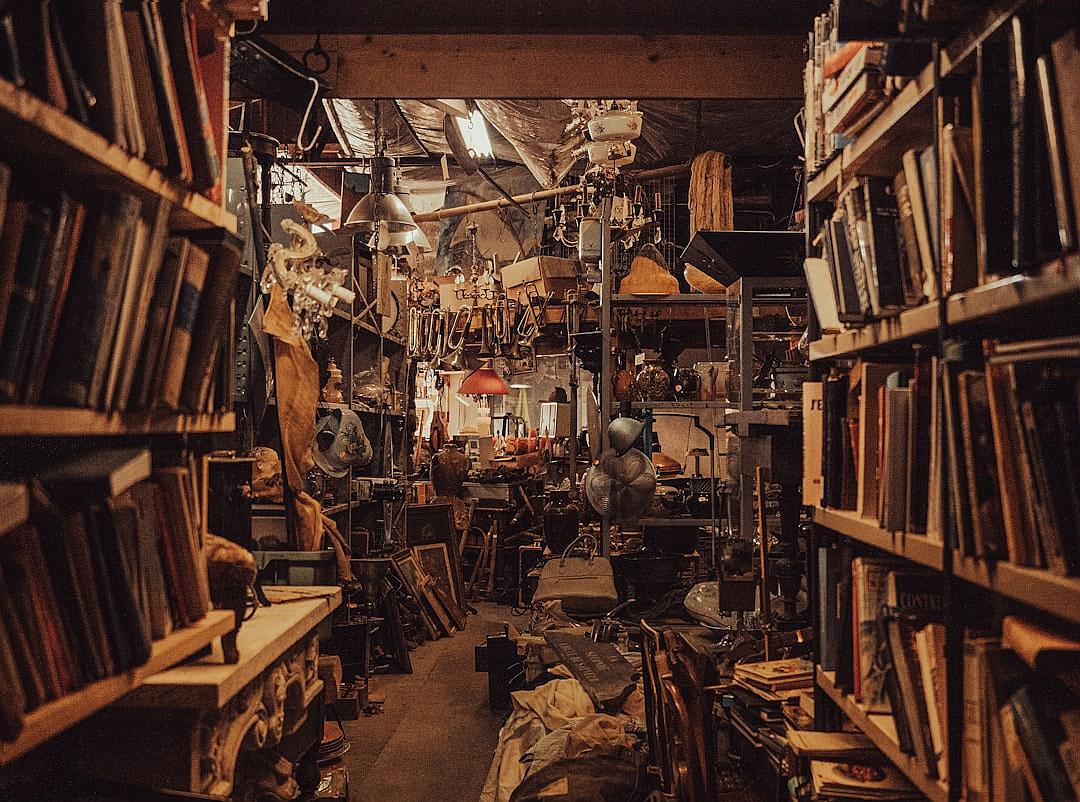 a photo of an old messy bookshop filled with books, trinkets and knickknacks. It’s dark inside the shop but there is light coming from above in some places. There’s lots of antique objects on shelves and hanging from ceiling. The atmosphere feels like it would be at night time. In one corner of the room we can see large boxes stacked up covered by black plastic sheeting. At dusk. Shot taken on Nikon Z9