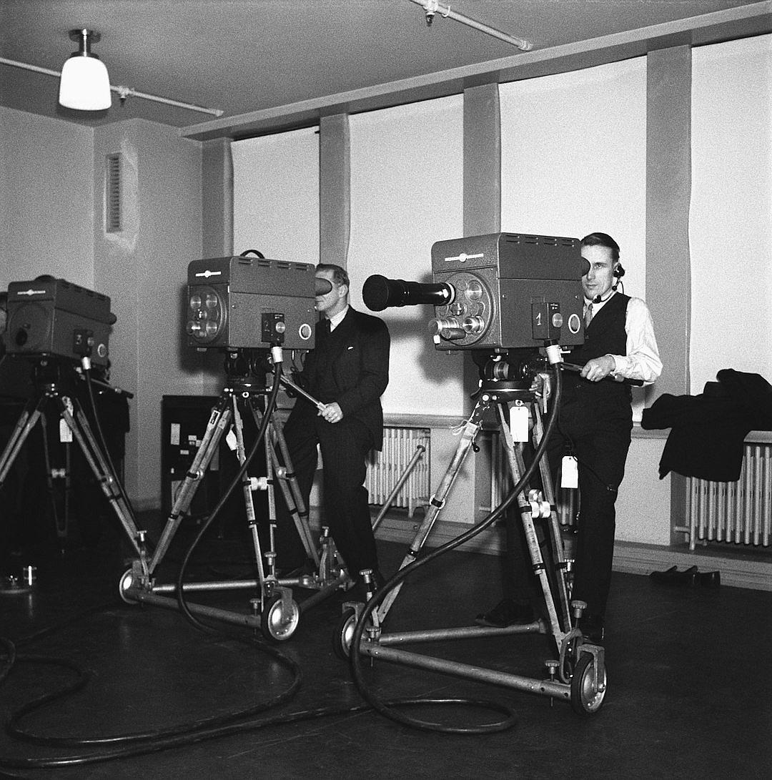 In the style of black and white documentary photography, an old photograph from around the year 2035 shows two men standing in front of cameras on tripods with large video cameras behind them inside a Swedish technical room. One man is wearing a formal suit while the other wears a casual outfit. The cameras are pointing towards a wall to capture some kind of sound or waves coming out.