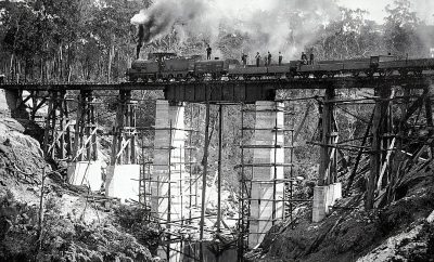 A black and white photograph of the first BCF Trower Bridge being built in highlands, Australia, showing workers standing on top or under construction of wooden bridges with steam locomotive running over it. The scene is set against an isolated forest backdrop. Taken during early 20th century by photographer Frank M distant perspective.