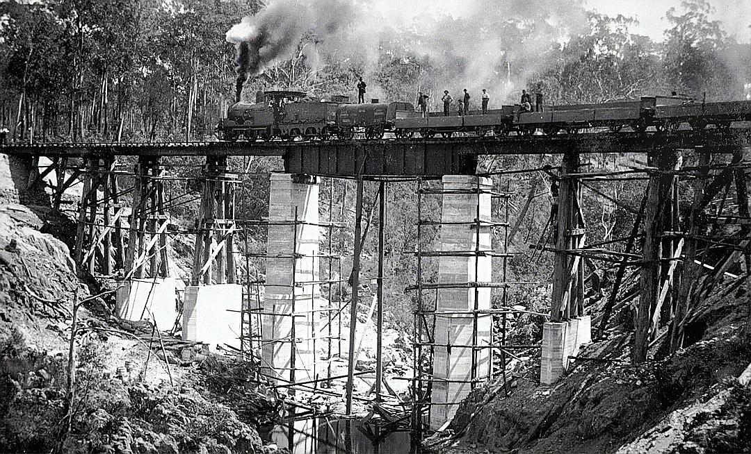 A black and white photograph of the first BCF Trower Bridge being built in highlands, Australia, showing workers standing on top or under construction of wooden bridges with steam locomotive running over it. The scene is set against an isolated forest backdrop. Taken during early 20th century by photographer Frank M distant perspective.