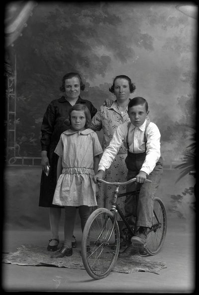 Old black and white photo of family with two kids, one girl in a dress standing next to a boy on a bike, posing for a portrait, in the studio style, from the 1920s, old photography, no background, vintage look, retro style.